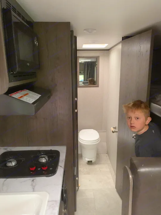 Curious boy peeking in an RV rental interior, showing a compact kitchen with stovetop and a small bathroom with toilet.. Jayco Jay Flight 2020
