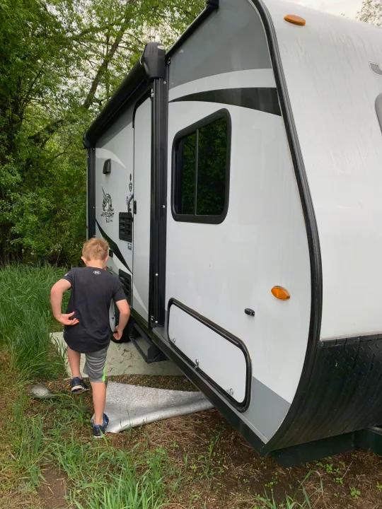 Child steps into an RV rental, a travel trailer with a black awning, amidst green grass and trees.. Jayco Jay Flight 2020