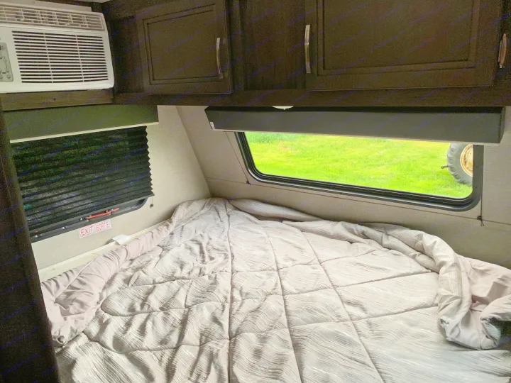 RV rental camper interior: cozy bed, AC unit, cabinets, and window with a view of green grass and a large tire outside.. Jayco Jay Flight 2020
