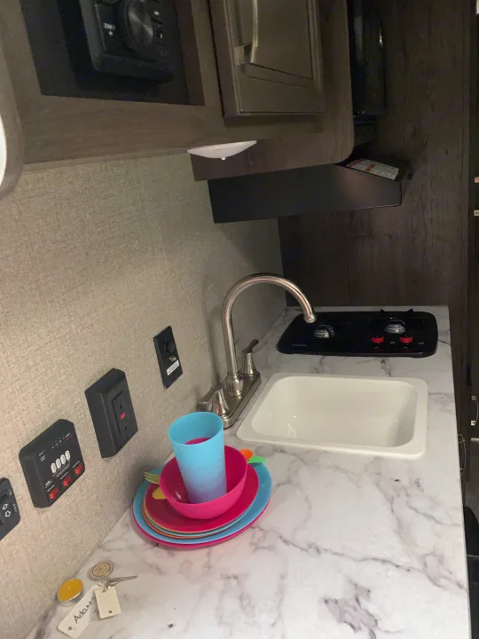 RV rental kitchen showing sink, two-burner stove, colorful dishes, and wall controls. Keys on marble counter.. Jayco Jay Flight 2020