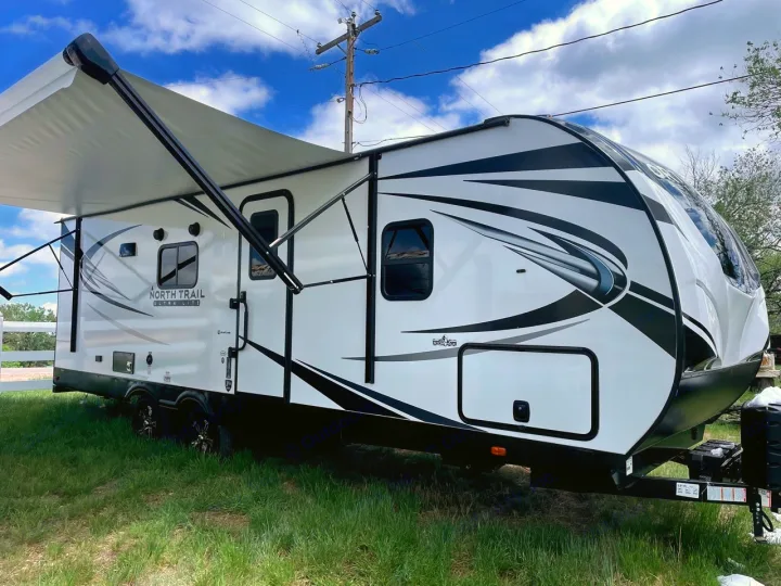 Bright white travel trailer with black graphics, awning extended, parked on grass. Perfect for an rv rental vacation!. Heartland RVs North Trail 2021