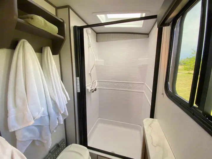 Well-lit RV rental bathroom featuring a shower stall, skylight, hanging towels, and outdoor view.. Cruiser RV M-277BHS 2021