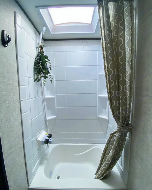 RV rental bathroom features a compact tub, shower, skylight, and a tied-back patterned curtain. Greenery hangs from the shower.. Gulf Stream Other 2021