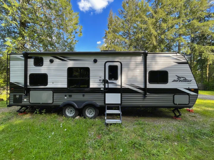 This grey Jay Flight by Jayco travel trailer rv rental is parked on grass amidst tall trees under a blue sky.. Jayco Jay Flight 2022