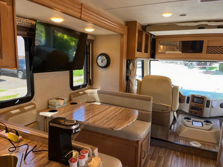 Modern RV rental interior, showing a dinette, kitchen with coffee maker, wall-mounted TV, and driver's cockpit.. Thor A.C.E. 2017