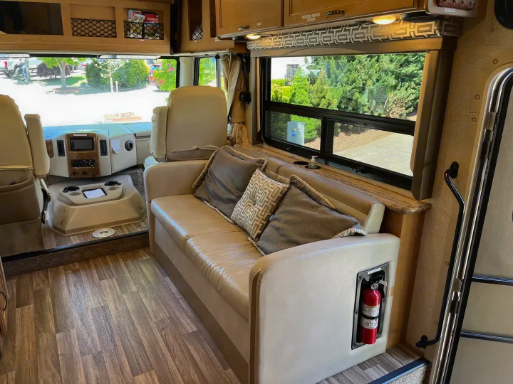 Inside a modern RV rental: comfortable living area with beige sofa, large windows, wood-look floor, and driver's cabin.. Thor A.C.E. 2017