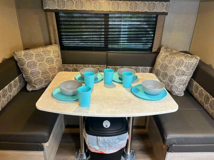 Modern RV rental dinette with black benches, patterned cushions, a light table set with blue/grey dishes, and a window with blinds.. Coachmen Catalina Summit Series 7 2022