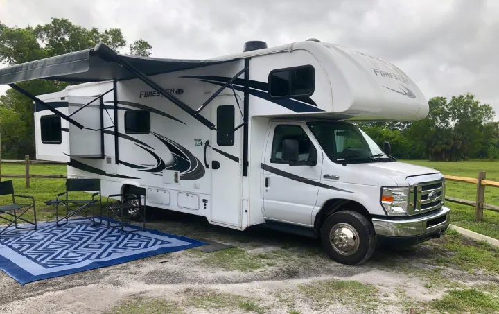 Class C RV rental parked at a serene campsite with an extended awning, chairs, and a blue outdoor mat.. Forester LE 2020