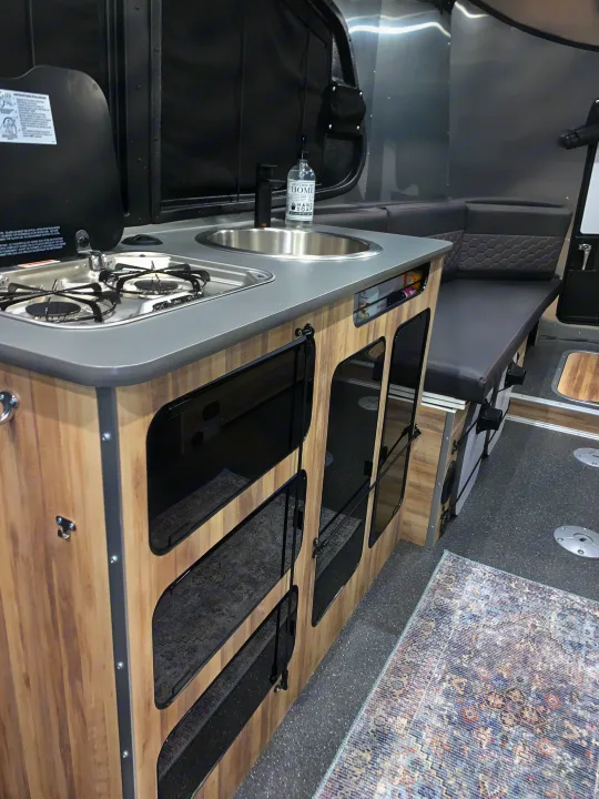Compact RV rental kitchen & living space: stove, sink, and bench seating with textured cushions.. Airstream Basecamp 2021