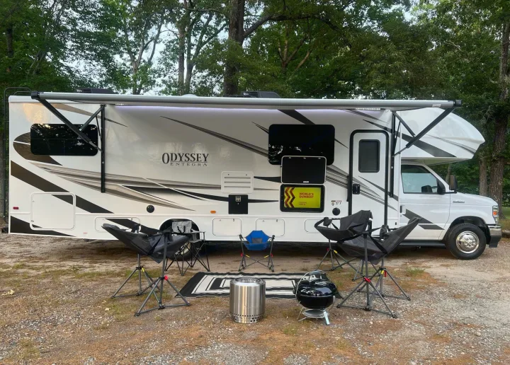 RV rental: A white Odyssey Entegra at a campsite. Awning out, with chairs, fire pit, and grill for relaxing.. Entegra Coach Odyssey 2022