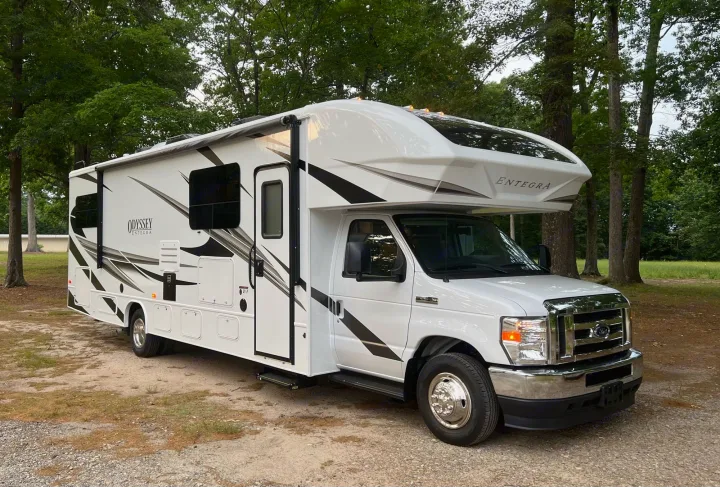 A sleek white Entegra Odyssey Class C rv rental, with black and gray graphics, parked on a gravel lot among trees.. Entegra Coach Odyssey 2022