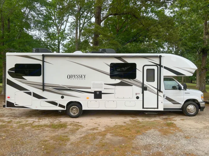 This white "Odyssey Entegra" Class C RV rental is parked on a dirt lot, surrounded by green trees.. Entegra Coach Odyssey 2022