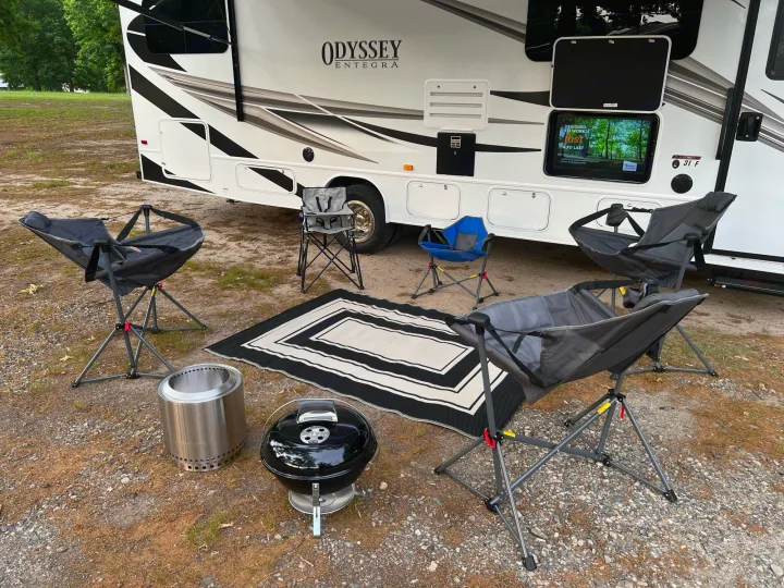 Relaxing RV rental campsite setup with outdoor TV, various camping chairs, Solo Stove fire pit, and Weber grill.. Entegra Coach Odyssey 2022