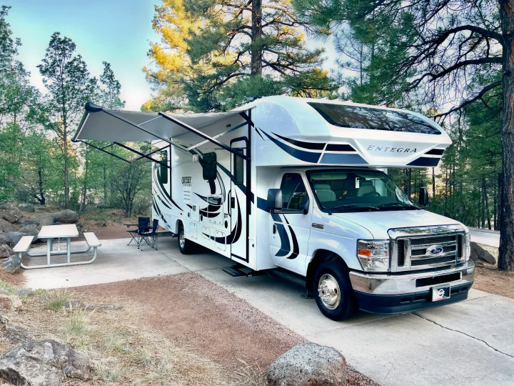 White Entegra Odyssey RV rental set up at a scenic wooded campsite with an awning, picnic table, and chairs.. Entegra Coach Odyssey 2021