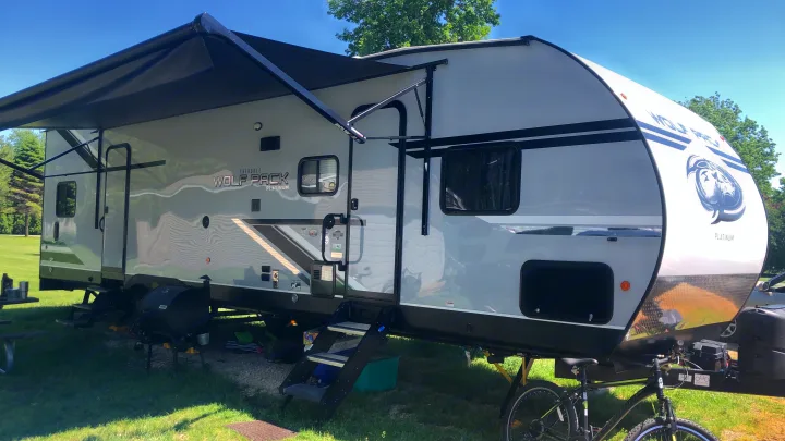 A spacious grey Wolf Pack Platinum RV rental is parked at a sunny campsite with its awning extended, perfect for outdoor living.. Forest River Cherokee Wolf Pack Toy Hauler 2020