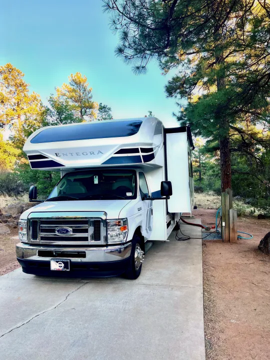A white Entegra Class C RV rental, hookups connected, is parked on concrete at a wooded campsite, ready for touring.. Entegra Coach Odyssey 2021