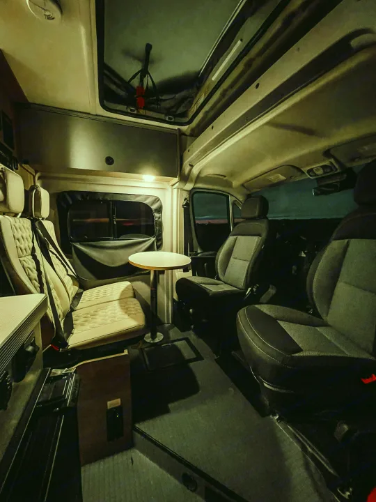 Well-lit RV rental interior at night. Features comfortable seating, a small table, and panoramic windows for a cozy travel experience.. Winnebago Solis 2022