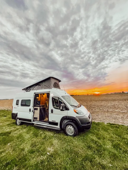 White camper van with pop-top & open door on green grass, beside a field at sunset. Ideal for an RV rental journey!. Winnebago Solis 2022