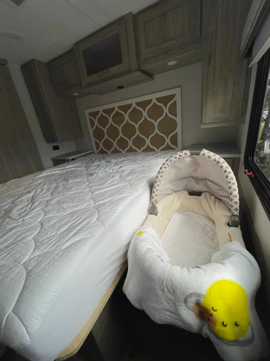 RV rental interior: A cozy bed, baby bassinet with chick blanket, and wooden storage cabinets create a family-friendly space.. Coachmen Pursuit 2021