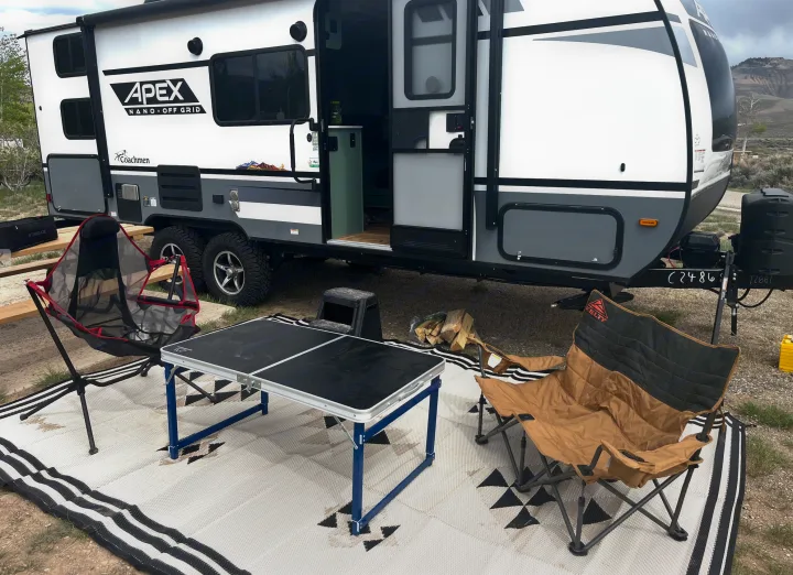 Enjoy this RV rental's outdoor camp setup: an Apex trailer with a folding table, two chairs, and mat on a natural site.. Forest River Coachmen Apex Nano Off-Grid 2022