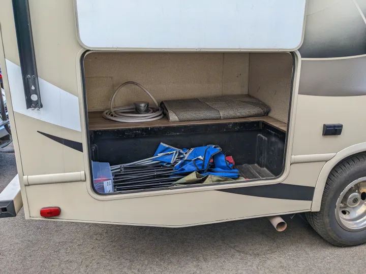 Open storage compartment of an RV rental with camping gear: folded chairs, hose, mat, and bucket inside.. Thor Motor Coach Four Winds 2017