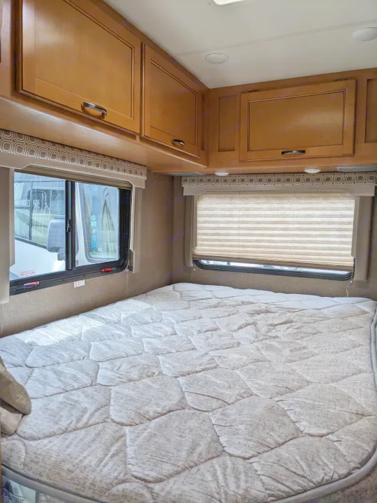 RV rental bedroom interior featuring a comfortable bed, overhead cabinets, and windows with patterned valances and shades.. Thor Motor Coach Four Winds 2017