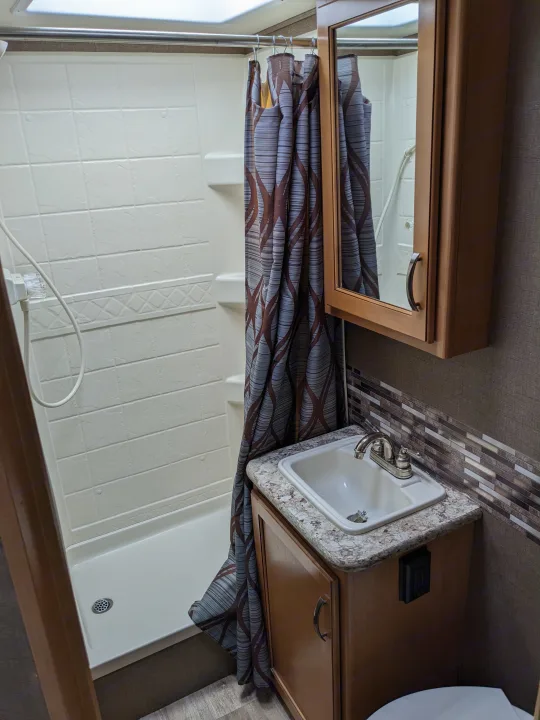 This compact RV rental bathroom features a functional shower with a patterned curtain and a small sink area with a mirrored cabinet.. Thor Motor Coach Four Winds 2017