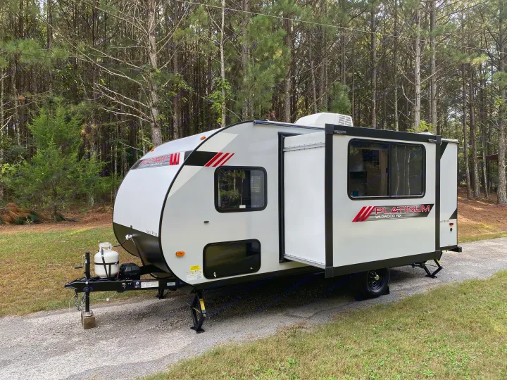 RV rental: White travel trailer with extended slide-out & propane tanks, parked on gravel in a forest setting.. Forest River Wildwood FSX Platinum 2021