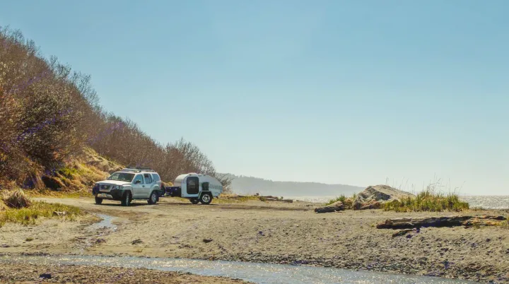 Silver SUV towing a white RV rental teardrop camper parked on a sandy beach by the ocean.. Little Guy Other 2012