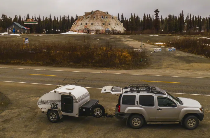 An RV rental setup: a silver SUV tows a teardrop camper. Parked by the road, with the abandoned Igloo Hotel in the background.. Little Guy Other 2012