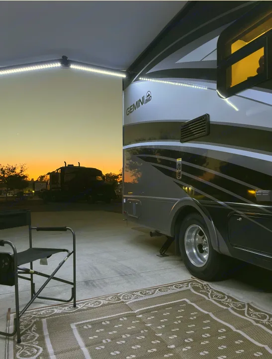 Serene dusk at an RV rental site, featuring a Thor Gemini motorhome with illuminated awning, cozy outdoor rug & chair setup.. Thor Gemini 2022