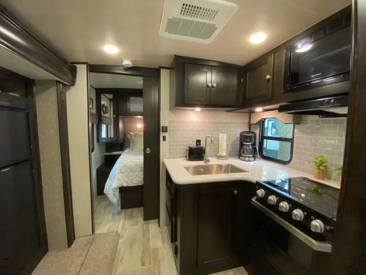 Experience comfort in this RV rental! Features a well-equipped kitchen with dark cabinets and a stove, opening to a cozy bedroom.. Heartland RVs North Trail 2019