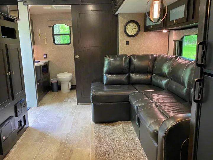 Spacious RV rental living area with a black leather sectional, wall-mounted TV, bathroom, and window view. Comfortable travel lodging.. Heartland RVs North Trail 2019