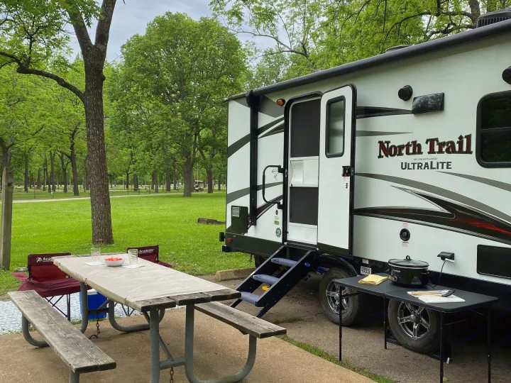 This RV rental campsite features a picnic table with a meal, chairs, and a slow cooker outdoors, set against a green park with trees.. Heartland RVs North Trail 2019