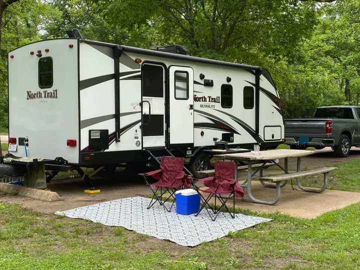 RV rental setup at a shaded campground: a white North Trail travel trailer, pickup truck, picnic table, and chairs.. Heartland RVs North Trail 2019