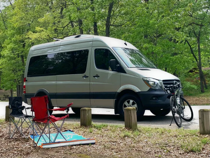 An RV rental camper van parked at a green campsite with bikes, two chairs, and a mat. Ideal for outdoor adventures.. Mercedes Benz 2017 Sprinter Van 2017