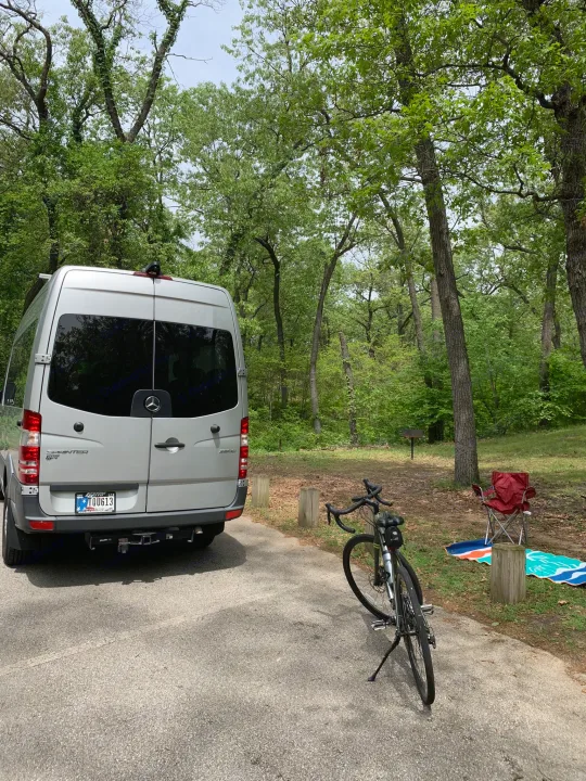 Silver RV rental van parked at a wooded campsite with a bike, red chair, and colorful mat.. Mercedes Benz 2017 Sprinter Van 2017