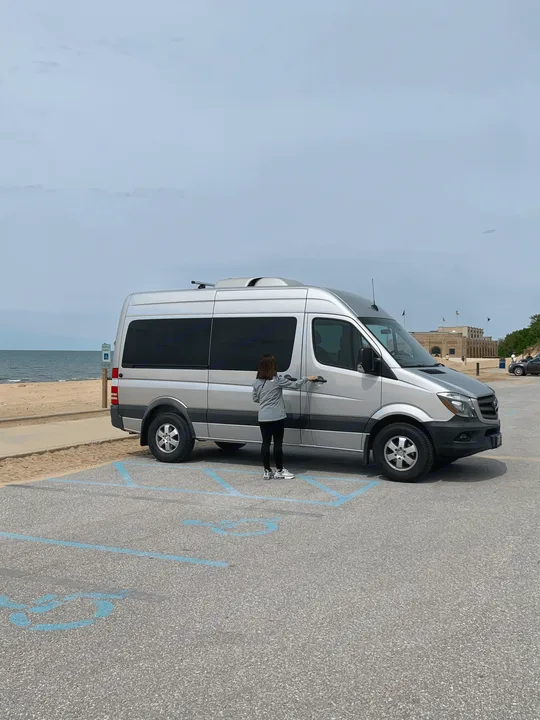 Woman opening a silver RV rental van in an accessible beach parking lot. Ideal for outdoor travel adventures.. Mercedes Benz 2017 Sprinter Van 2017