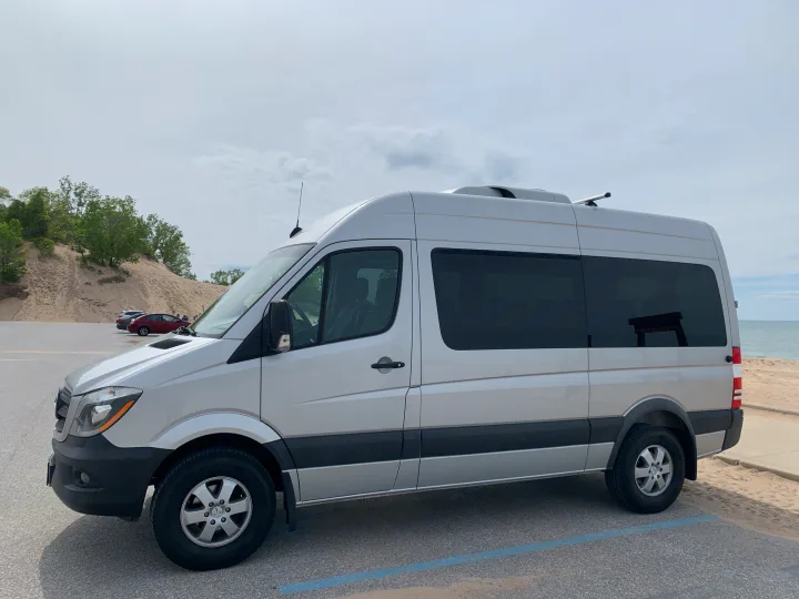 Silver Sprinter camper van (potential RV rental) parked by sandy dunes and a beach under a cloudy sky.. Mercedes Benz 2017 Sprinter Van 2017