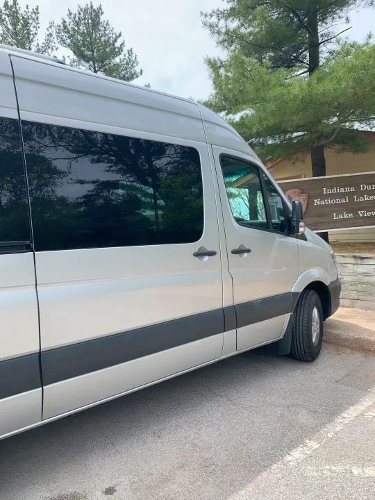 Silver RV rental van parked at Indiana Dunes National Lakeshore, surrounded by pine trees.. Mercedes Benz 2017 Sprinter Van 2017