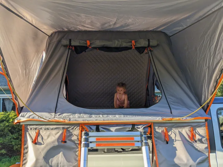 A happy toddler peeks from a grey rooftop tent mounted on a vehicle, accessible by a ladder. Perfect for RV rental trips!. Jeep gladiator overland Jeep gladiator overland 2020