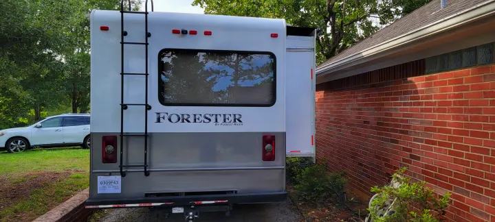This white & grey Forester RV rental, with a ladder & large rear window, is perfectly parked beside a brick house.. Forest River 3171DS 2016