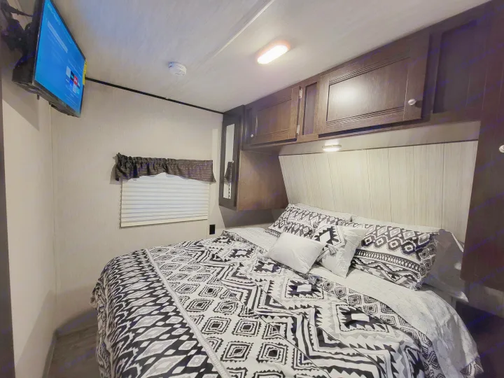 RV rental bedroom: Interior shows a bed with black/white geometric bedding, a wall-mounted TV, and dark wood cabinets.. Heartland RVs Pioneer 2019