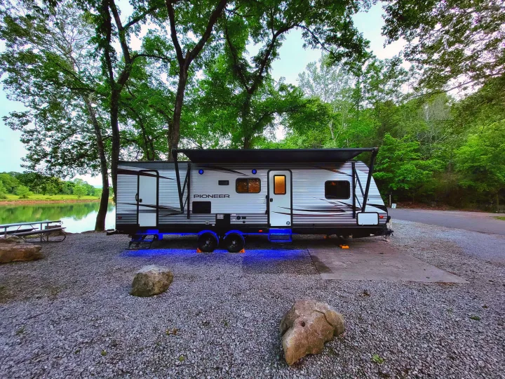 Lakeside travel trailer RV rental with blue underglow at a gravel campsite, offering a serene outdoor escape.. Heartland RVs Pioneer 2019