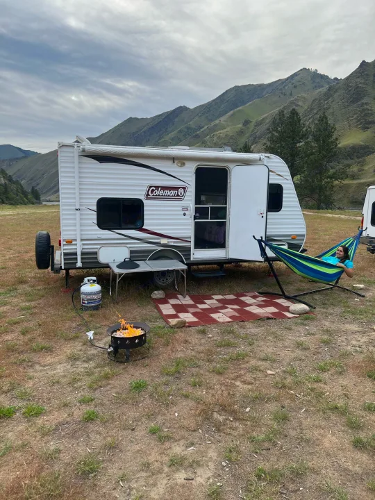 A person relaxes in a hammock beside a white travel trailer (RV rental) with a lit fire pit and mountains.. Dutchmen Coleman Expedition LT 2013
