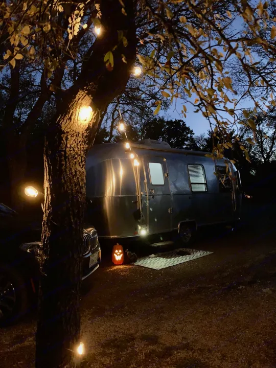 Illuminated Airstream RV rental at dusk, decorated with string lights & a jack-o'-lantern, offering a festive camping experience.. Airstream Sport 2019