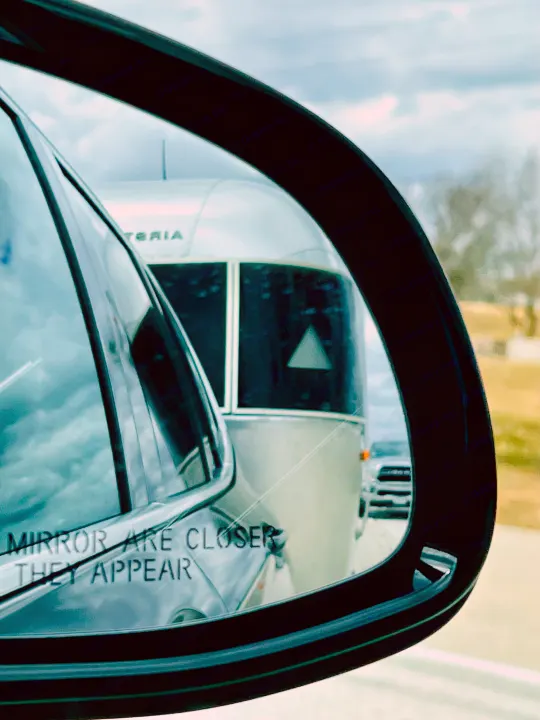 An Airstream RV rental is reflected in a car's side mirror. Text reads 'MIRROR ARE CLOSER THEY APPEAR', during travel.. Airstream Sport 2019