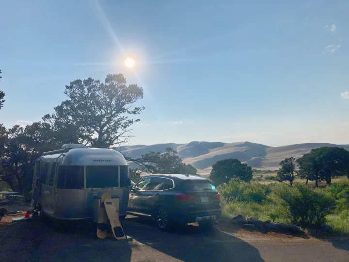 A sleek RV rental (Airstream) and SUV parked at a desert campsite, with towering sand dunes nearby.. Airstream Sport 2019