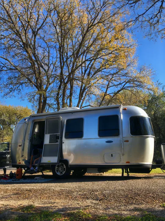 Airstream rv rental at a sunny campsite. Person inside open door, trees above with sparse yellow leaves.. Airstream Sport 2019