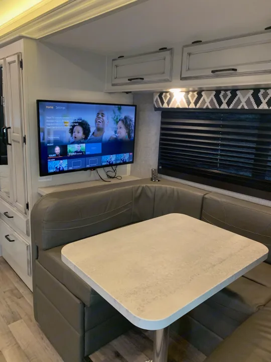 Cozy RV rental interior: a U-shaped dinette with a light-colored table, and a wall-mounted TV showing channel options.. Entegra Odyssey 2022